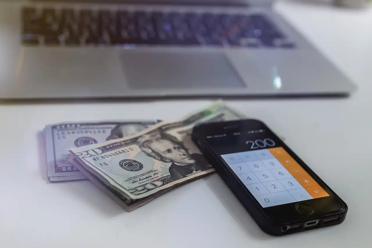 A calculator sits atop some cash on a desk next to a computer