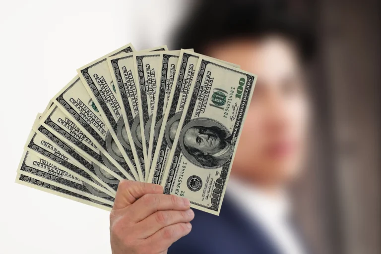 A man holds $100 bills, fanned out, in front of him while he stays out of focus in the background