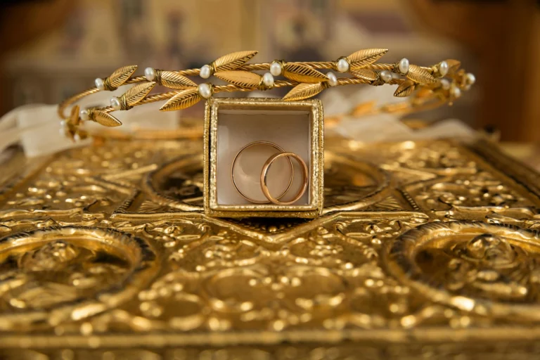Two gold wedding bands are nestled in a small jewelry box atop an ornate gold tabletop. A gold leaf and freshwater pearl headpiece rests decoratively on top of the box.