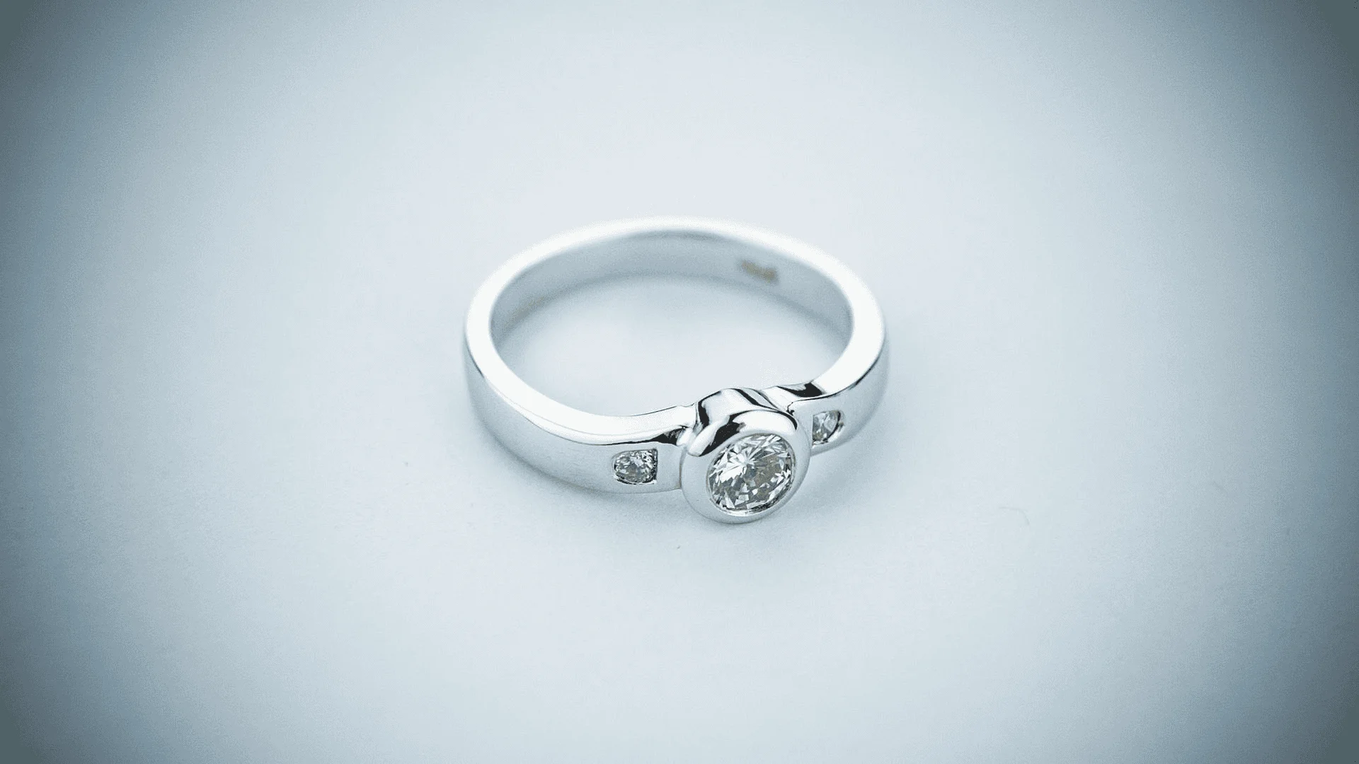 A white gold ring features a bezel set round cut center stone, flanked by inlaid diamonds on each side